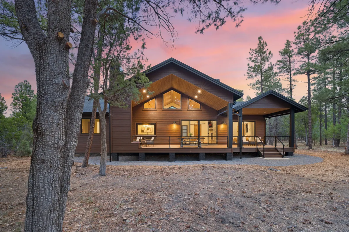 A spacious wooden deck extends from the home, framed by large windows and surrounded by tall trees. The sunset sky casts a warm glow over the tranquil landscape, suggesting a seamless blend between indoor comfort and the natural world outside.