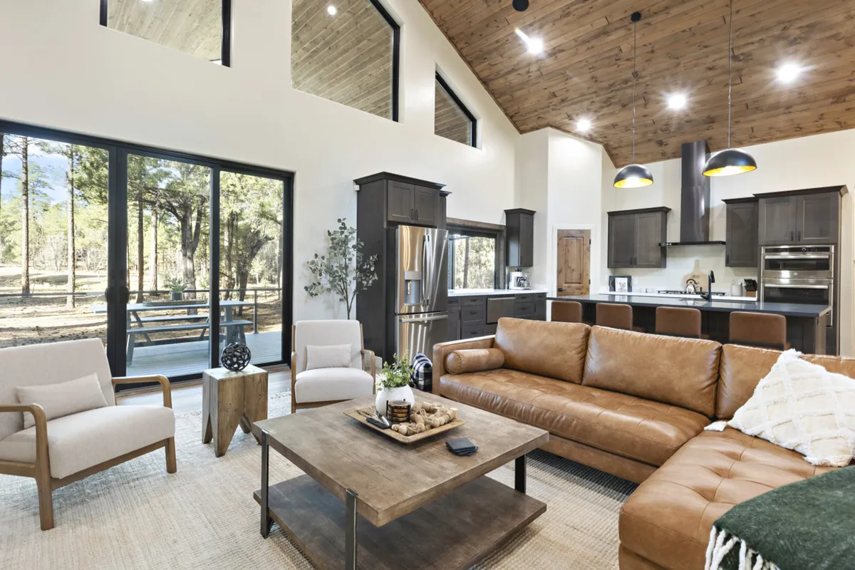 An inviting living space featuring a large leather sofa and a sturdy wooden coffee table, with the kitchen area visible in the background. Expansive floor-to-ceiling windows flood the space with natural light and offer serene sunset views. The kitchen area is subtly highlighted, showing its proximity to the living area and reinforcing the concept of an open, connected communal space.