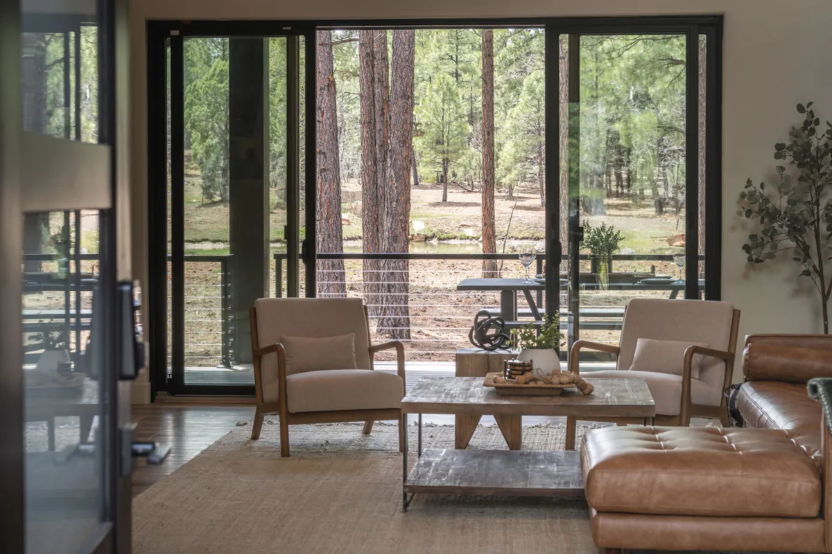 A spacious great room with large glass doors opening to natural greenery, featuring a wooden table for shared meals, comfortable seating areas for conversation or games, and plenty of light. The scene feels tranquil and welcoming, with a seamless connection between indoors and the lush outdoors.