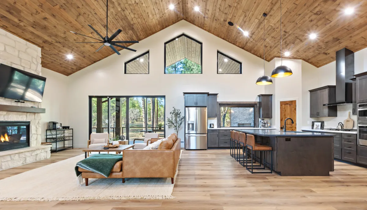 This image showcases a luminous great room with soaring vaulted ceilings, expansive windows framing lush forest views, a cozy fireplace, and an inviting island kitchen nearby. A comfortable sofa anchors the sitting area, creating a welcoming space perfect for gatherings and relaxation.
