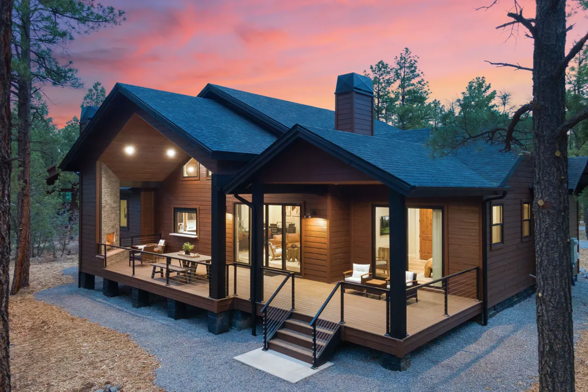 This image shows the front porch of Hidden Pond Retreat, framed by large windows and wooden siding, surrounded by towering trees beneath a glowing sunset sky. The inviting porch hints at opportunities for outdoor relaxation, dining, or enjoying a refreshing break in nature during the workday.