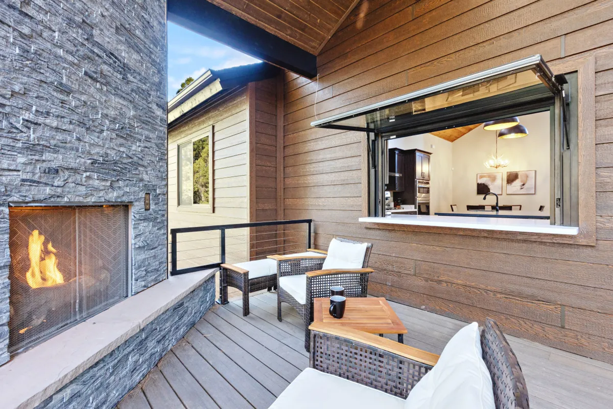 An inviting outdoor seating area arranged around a fireplace, with an open window connecting the space to the indoors. The area overlooks natural surroundings, ideal for group gatherings and outdoor relaxation. The lighting suggests a cozy atmosphere perfect for sunset views or evening get-togethers.
