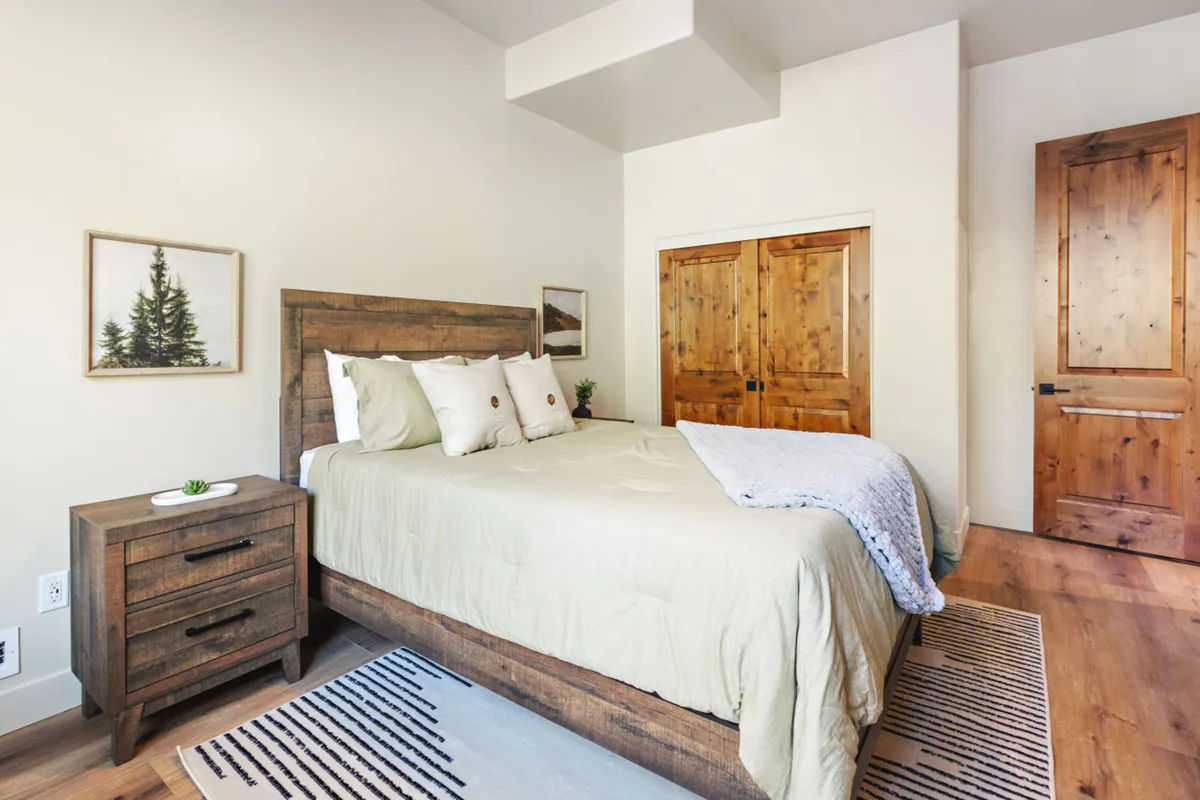 A calm bedroom with a well-made bed, nightstand, wooden doors, and framed artwork. Soft natural light filters through the window, creating a cozy setting ideal for relaxation and restful nights.