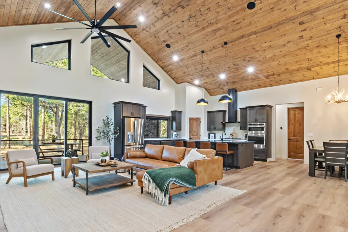The image highlights a stylish interior with modern leather sofas arranged for socializing, large windows offering scenic forest views, and a wooden ceiling that adds warmth. The kitchen island and open dining area in the background reinforce the sense of a communal living space.