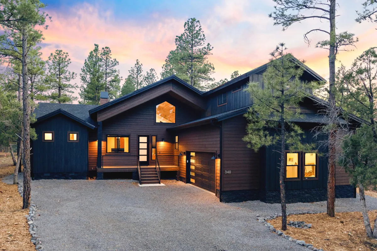 The image showcases the modern architecture of a spacious single-family house nestled among tall pine trees. Warm, inviting light glows from the expansive windows, illuminating the welcoming porch—an ideal spot for gathering with family. The tranquil surroundings and natural beauty of the pine forest set a serene mood, hinting at opportunities for sunset views and nature walks just steps away.