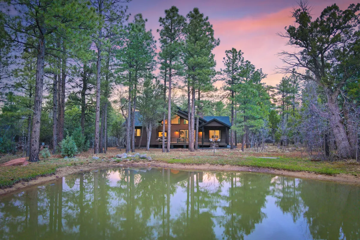 An exterior scene highlighting the house, a tranquil pond, and surrounding trees. The setting evokes a peaceful retreat, with the pond reflecting the sunset and trees adding a sense of seclusion and natural beauty.