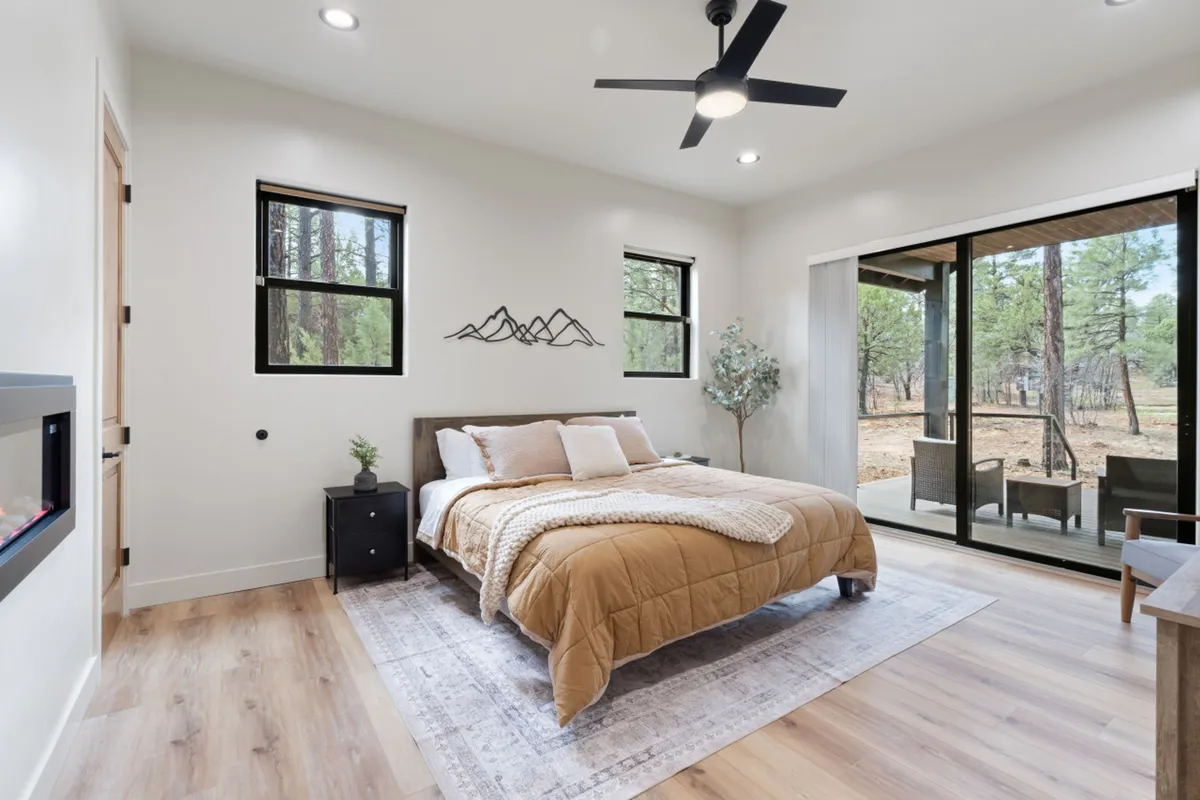 A serene bedroom featuring a comfortable bed with mountain-inspired decor, large windows that offer views of the surrounding nature, and an outdoor seating area just beyond the glass. The atmosphere is cozy and tranquil, inviting relaxation and restful sleep.