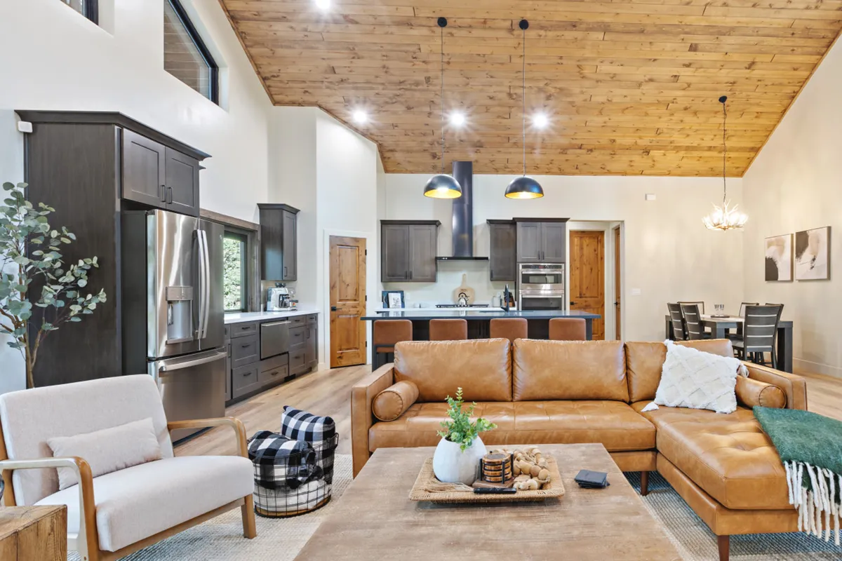 This inviting interior scene centers on a large leather sectional sofa and a central coffee table, with the kitchen island clearly visible in the background. The open concept space flows naturally from the living area to the kitchen, allowing easy interaction between those cooking and guests or family members relaxing nearby. Warm wood tones and ambient lighting add a rustic, homey charm, while modern appliances and finishes suggest practical convenience.