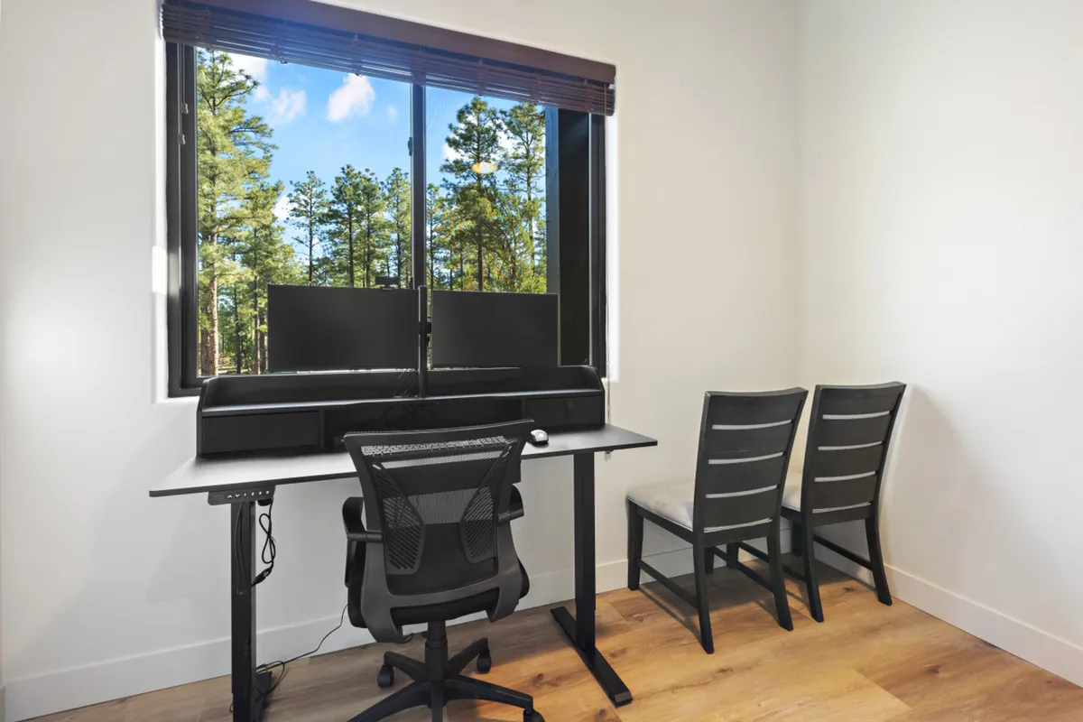 A bright, dedicated office space featuring a large window framing a lush forest view. The room includes a modern desk with dual monitors and a comfortable office chair, showcasing an inviting setup for productivity while being surrounded by nature.
